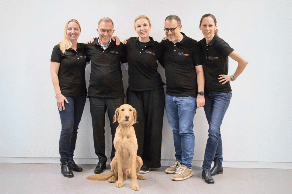 Fünf Personen in schwarzen SUST-Poloshirts stehen Arm in Arm vor einer weissen Wand, vor ihnen sitzt ein hellbrauner Hund