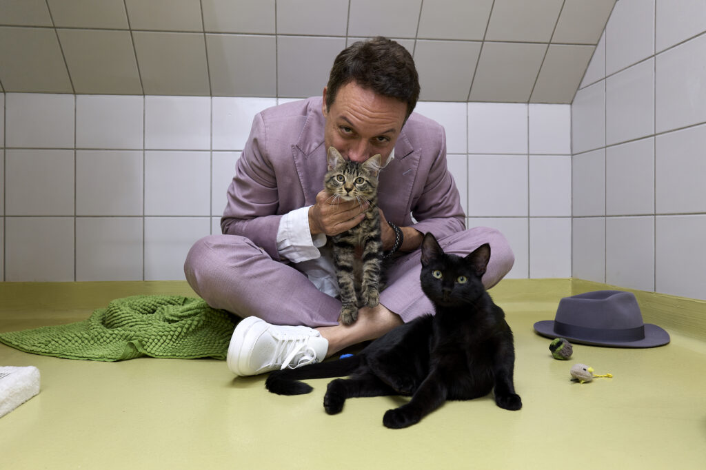 Camen sitzt im Katzenzimmer eines Tierheims am Boden, hält ein getigertes Kätzchen, vor ihm liegt eine schwarze Katze
