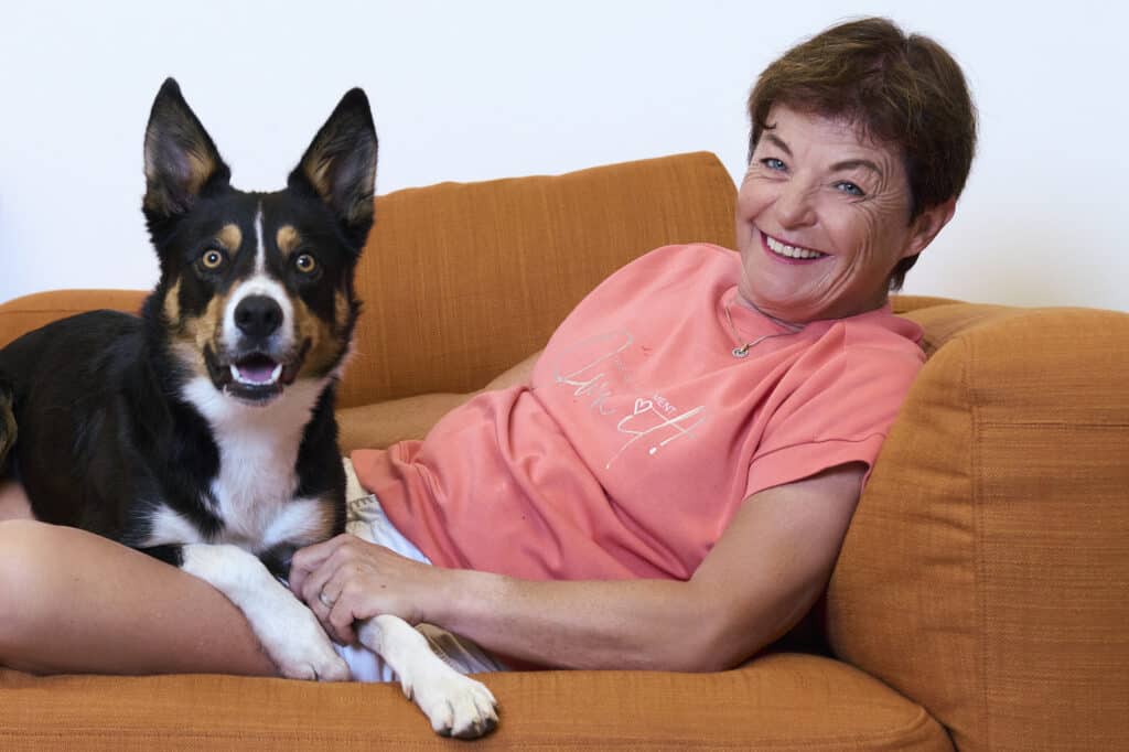 Monika Fasnacht liegt lachend auf einem orangen Sofa, vor ihr ihr Border Collie